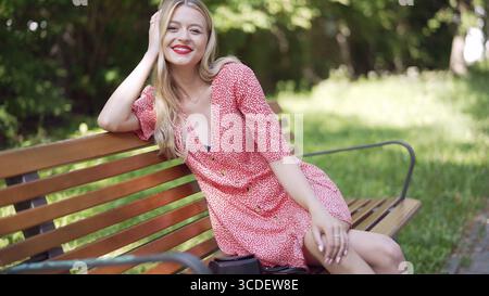 Una giovane donna gioiosa con capelli biondi e rossetto rosso gode di una giornata di sole, seduta su una panchina del parco, trasuda felicità e positività Foto Stock
