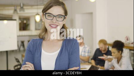 Ritratto di una giovane bruna che indossa abiti formali e occhiali da vista neri che posa con le mani incrociate e sorride alla macchina fotografica in ufficio Foto Stock