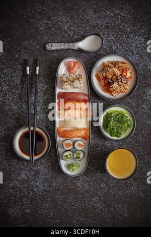 Assortimento di piatti asiatici. Vari sushi roll posizionati su piatti di ceramica. Insalate kimchi e goma wakame. Zuppa di soia e bacchette ai lati. bac scuro e grungy Foto Stock