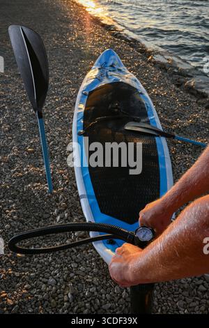 Uomo che gonfia la tavola sup con una pompa a mano sulla spiaggia di ciottoli al tramonto, preparandosi per gli sport acquatici. Foto Stock
