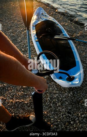 Sportsman che gonfia il sup board con pompa a mano sulla spiaggia di ciottoli al tramonto, preparandosi per il paddleboard. Foto Stock