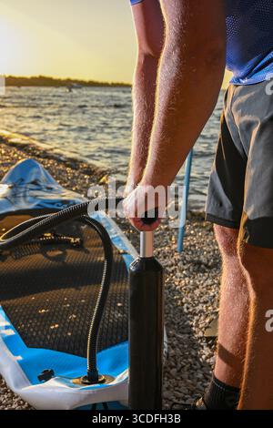 Uomo che gonfia la tavola sup con una pompa a mano su una spiaggia di ciottoli al tramonto. Foto Stock