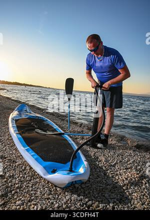 Sportsman che gonfia il sup board con pompa manuale sulla spiaggia di ciottoli al tramonto, preparandosi per il paddleboard. Foto Stock