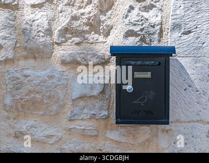 Moderna cassetta postale nera con una grafica decorativa a forma di uccello montata su un antico muro in pietra rustica in piena luce del sole. Contrasto tra vecchio e nuovo Foto Stock