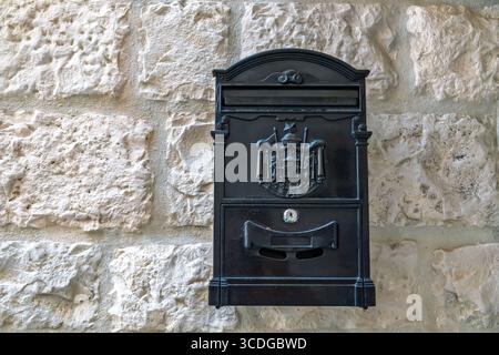 Una cassetta postale nera ornata con uno stemma, montata su un antico muro di pietra rustica. Cassetta postale in metallo nero vintage sull'antico muro di pietra di Foto Stock