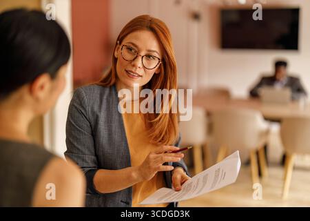 Donna d'affari sicura di sé in occhiali che discute documenti con una collega in ufficio Foto Stock