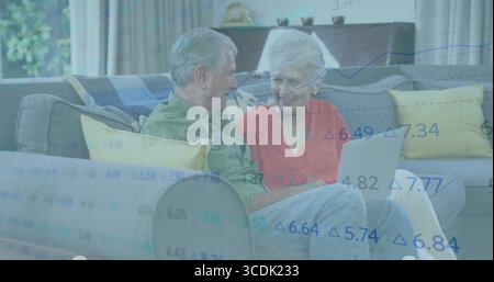 Una coppia senior che esamina i dati delle scorte su un notebook color argento nel soggiorno, con cuscini gialli Foto Stock