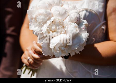 Un primo piano cattura le mani delle spose con delicatezza tenendo in mano un bouquet lussureggiante di peonie bianche, simboleggiando l'amore e i nuovi inizi, immersi nel caldo sole Foto Stock