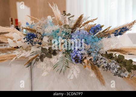 Un lungo corridore da tavolo mostra uno straordinario centro nuziale, con erba di pampas, delicate ortensie blu e fogliame di eucalipto in un boh neutro Foto Stock
