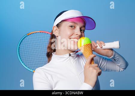 Giovane atleta in abbigliamento sportivo bianco con racchetta e cono che reggono la palla su sfondo blu da studio, divertente concetto di sport estivo per gli annunci Foto Stock