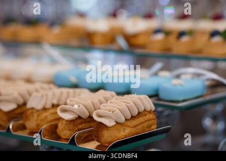 Uno scatto ravvicinato mostra file di delicati bocconcini di panna con glassa beige chiara e macaron blu pastello disposti su vassoi di servizio a più livelli, allettanti Foto Stock