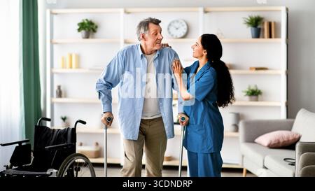 Infermiera femminile che aiuta i maschi anziani a camminare con le stampelle a casa. Assistenza medica professionale per pazienti anziani disabili Foto Stock
