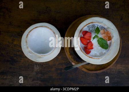 Un'insalata di frutta mista, un dessert colorato in mezza noce di cocco fresco, su un tavolo di legno scuro. Foto Stock