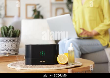 Umidificatore d'aria con olio essenziale e limoni sul tavolo contro la donna che usa il laptop a casa, primo piano Foto Stock