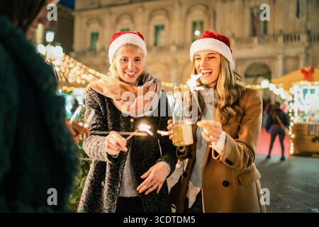 Gruppo di giovani amici felici che ridono e festeggiano con scintillanti e champagne in un mercatino di Natale all'aperto, visto da dietro le spalle. Foto Stock