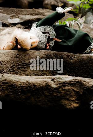 Una bambola con i capelli biondi e un vestito verde è adagiata su una superficie di pietra. Foto Stock