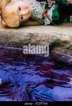 Una bambola con i capelli biondi e gli occhi azzurri è adagiata su una superficie di pietra accanto a un corpo d'acqua. L'acqua ha una superficie scura e riflettente. Foto Stock
