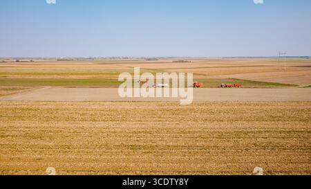 Sopra la visuale, la trebbiatrice mentre passa tra i campi agricoli su un sentiero rurale, viaggia su una strada polverosa. Due trattori con rimorchi vengono azionati sullo stesso modo Foto Stock