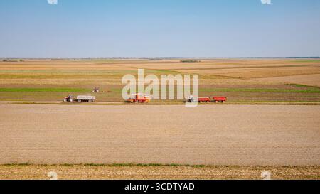 Sopra la visuale, la trebbiatrice mentre passa tra i campi agricoli su un sentiero rurale, viaggia su una strada polverosa. Due trattori con rimorchi vengono azionati sullo stesso modo Foto Stock
