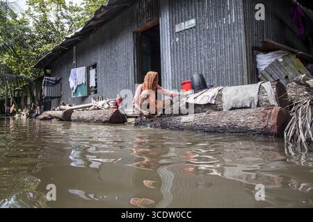 Noakhali, Bangladesh - 7 settembre 2024: Vista di una donna arroccata su tronchi in mezzo alle acque alluvionali, il suo scialle arancione che rispecchia i toni tenui del paesaggio sommerso, accanto a una casa in metallo ondulato. Foto Stock