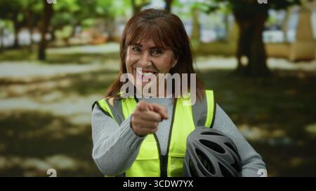 Una donna anziana ispanica sorride in un parco mentre punta la macchina fotografica, indossa un giubbotto riflettente e tiene in mano un casco da bicicletta, incarnando la sicurezza durante un Foto Stock