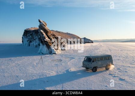 La vista aerea di un aspro affioramento roccioso con formazioni ghiacciate contrasta con la vasta distesa innevata dove un furgone attraversa il paesaggio ghiacciato, il lago Baikal, Russia. Foto Stock
