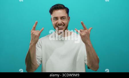 Il giovane con la barba che fa gesti di roccia sorride con fiducia su uno sfondo blu vibrante, mostrando energia e giovinezza. Foto Stock