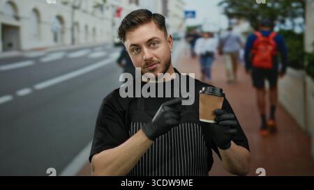 Il giovane con la barba nel grembiule punta alla tazza di caffè mentre si trova in strada, evidenziando il suo ruolo di cameriere con i guanti all'aperto in una città Foto Stock