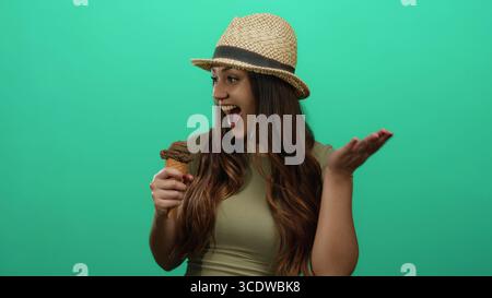 Donna che si gusta un gelato su uno sfondo verde, indossa un cappello e sorride, ritraendo un momento vivace con un dessert al cioccolato. Foto Stock