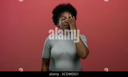 Donna che ride con la mano sul viso in piedi su sfondo rosso isolato, che esprime gioia e sorpresa, acconciatura afro, camicia grigia, giovane Am africana Foto Stock