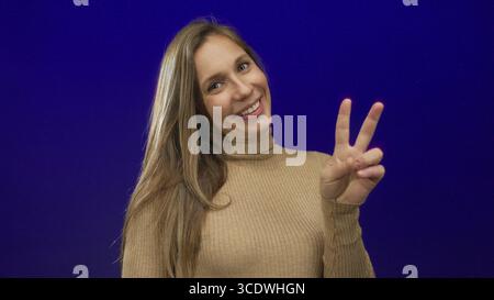 Donna sorridente e che fa un segno di pace davanti a uno sfondo blu che mostra fiducia e positività. Foto Stock