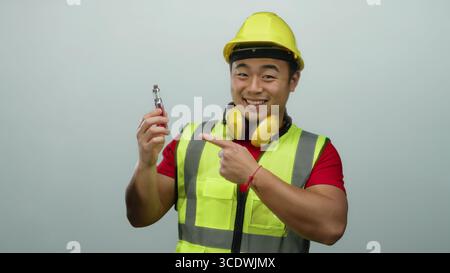 Un giovane uomo che indossa attrezzatura da costruzione e un elmetto su sfondo bianco mostra un dispositivo vape con un sorriso sicuro e un gesto che suggerisce il pollice Foto Stock