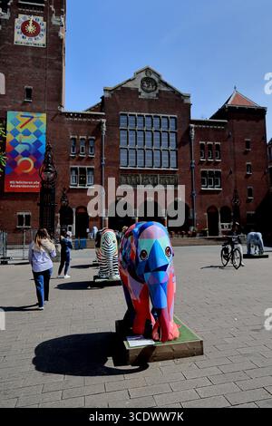 Sculture colorate di elefanti dipinte su Beursplein, Amsterdam, Paesi Bassi, sorgono nella piazza con pedoni e una facciata storica in luce soffusa. Foto Stock