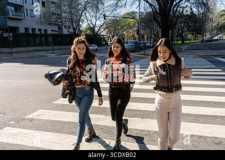 Tre studentesse stanno camminando e chiacchierando su un passaggio pedonale in città durante una giornata di sole Foto Stock