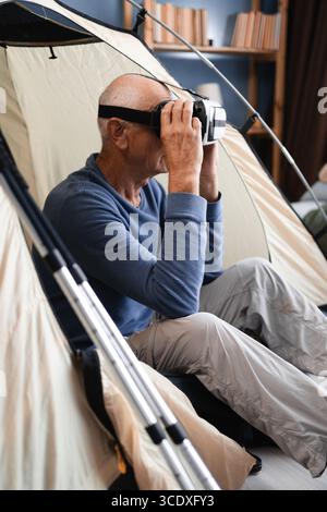 L'anziano seduto nella tenda del campeggio si diverte a giocare con gli occhiali per la realtà virtuale a casa al chiuso. Foto Stock
