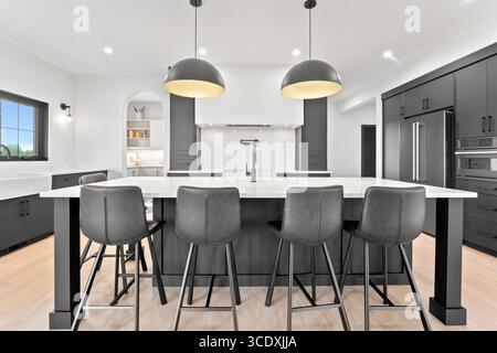 La cucina è spaziosa e ben illuminata, con una grande isola al centro e quattro sedie nere che la circondano. Le sedie sono posizionate in una diversa posizione Foto Stock