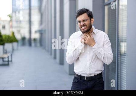 Un uomo in abbigliamento formale sta fuori tenendo la gola, mostrando disagio e dolore. Sembra che stia vivendo un problema medico legato alla sua gola in un ambiente urbano. Foto Stock