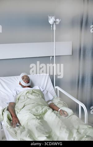 Attraverso le persiane colpo di giovane afroamericano che trascorre giorni in ospedale dopo ferita alla testa dormendo mentre riceve la terapia IV Foto Stock