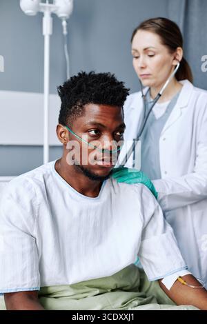 Scatto selettivo verticale di un giovane medico caucasico adulto che ascolta i suoni interni di cuore e polmoni dei pazienti neri utilizzando lo stetoscopio Foto Stock