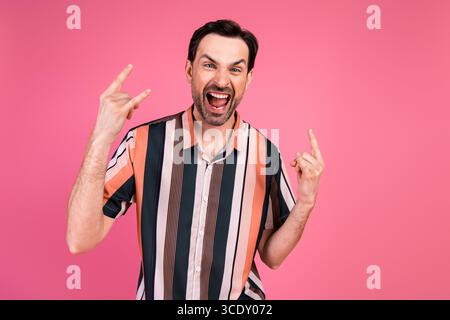 Giovane giocoso che fa gesti rock and roll su sfondo rosa e indossa una colorata camicia a righe Foto Stock