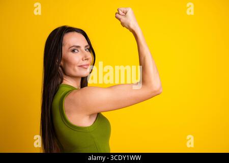 Giovane donna sicura di sé dallo stile casual che si flette il braccio con un sorriso su un vivace sfondo giallo Foto Stock
