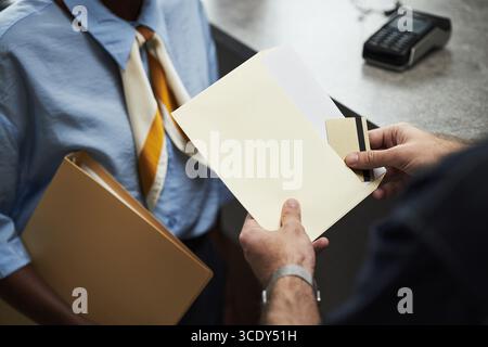 Le mani di un uomo che consegna la busta e prende la carta di credito, mentre sono servite da Black Woman che tiene la cartella, entrambe in banca vicino al terminale di pagamento, non sono visibili Foto Stock