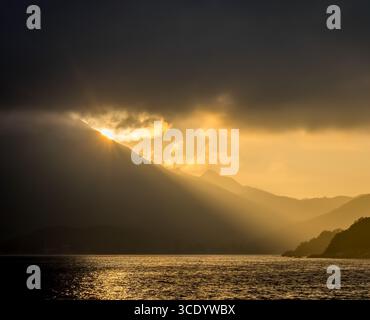 Sole d'oro sotto un drammatico cielo buio che tramonta sulle montagne di Lantau, Hong Kong, gettando pozzi di luce sulla valle, sulla costa e sul mare sottostante. Foto Stock