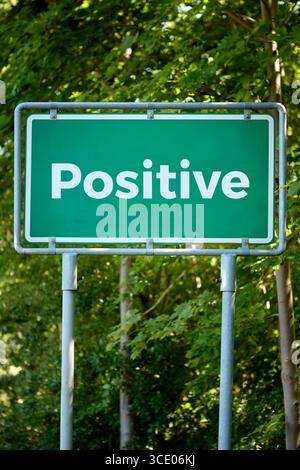 Un cartello stradale verde in stile autostrada che mostra la parola positivo in grassetto bianco. Adagiato all'aperto contro lussureggianti alberi verdi, simboleggia la positività e la gioia Foto Stock