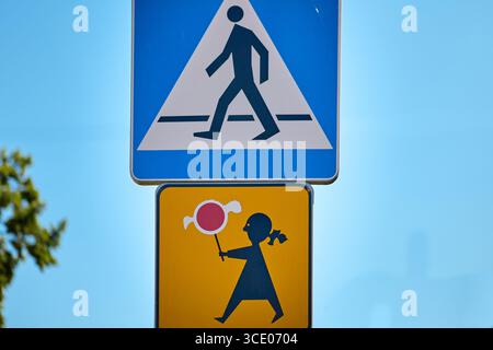 Un cartello blu di attraversamento pedonale sopra un cartello di avvertimento giallo che raffigura un bambino con in mano un lecca lecca. Indica un'area di attraversamento vicino a una scuola o a un playgroun Foto Stock