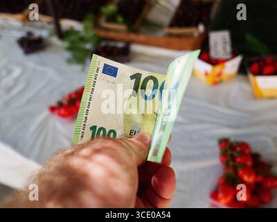 Primo piano di una mano che regge una banconota da 100 euro in un mercato all'aperto di Maiorca con sfondo sfocato di prodotti freschi Foto Stock