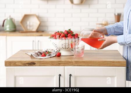 Donna con fragole fresche in colino che versano succo di fragole in vetro su un tavolo di legno in cucina Foto Stock