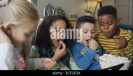 Mangiare, popcorn e bambini con tablet, casa e guardare cartoni animati con gli amici e intrattenimento. Rilassanti, gruppi e bambini con snack per film Foto Stock
