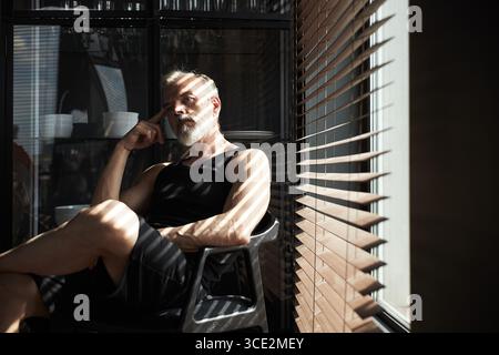 Uomo anziano caucasico seduto con cura all'interno vicino alle tende della finestra Foto Stock