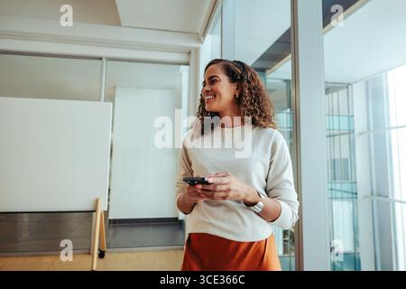 Una donna sorridente si trova in un luminoso e moderno studio d'ufficio, con in mano uno smartphone. Temi della tecnologia, dell'innovazione e della professionalità. Foto Stock